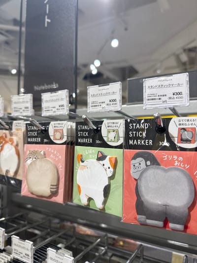 This image presents a detailed view of a retail display rack within a store in Kawanishi, Japan, near coordinates 34.826 latitude and 135.412 longitude. The brightly lit interior features a metal display unit showcasing multiple packages of "Stand Stick Markers," a type of sticky note. Each product package is individually sealed in clear plastic and hangs from a metal hook. The sticky notes inside are shaped as the hindquarters of various animals, specifically identifiable as a striped cat, a white-and-calico cat, and a gray gorilla. Each package indicates "30枚入" (30 sheets included) and is labeled with Japanese text describing the animal, such as "ゴリラのおしり" (Gorilla's butt) for the gorilla design. Transparent acrylic price holders are positioned above each product, displaying the brand "マインドウェイブ" (Mind Wave), the product name "スタンドスティックマーカー," a base price of "¥300," and a tax-inclusive price of "(税込 ¥330)." The "Loft" retailer logo is also visible on some price tags. In the blurred background, a sign partially visible above the display reads "Notebooks" in English and Japanese, indicating a stationery section. No people are visible in the scene.