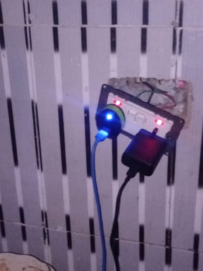 The image shows a close-up of a wall with a light switch and electrical outlets in Pantinapu, Nigeria. Two red indicator lights are illuminated on the switch plate. A blue LED light is visible on a charging device plugged into one of the outlets. Another black charging device with a red indicator light is also plugged into the switch plate. A blue cable extends from the charging device with the blue LED. The wall appears to be tiled with a vertical striped pattern in shades of grey and white.
