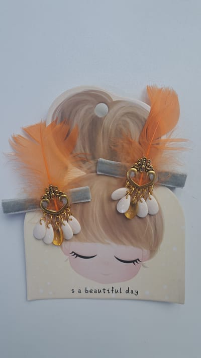 The image displays two hair clips, each featuring an orange feather, a grey velvet ribbon, and a gold-colored charm with white and yellow pendant beads. The hair clips are attached to a card depicting a cartoon girl with closed eyes and the text "s a beautiful day." The background is a plain, light-colored surface. There is no indication of the city of Altai, Kazakhstan, in the image.