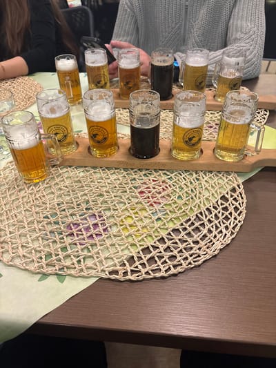 A collection of small beer mugs, some filled with dark beer, arranged on a wooden tray. The mugs are branded with "PIVO SNEZKA OSADNA". The tray is set upon a circular woven placemat with a floral patterned cloth underneath. A wooden table surface is visible to the right and parts of people are visible on the left side.