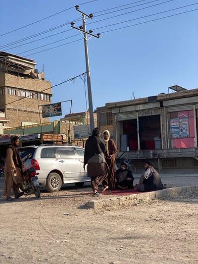 The image displays an outdoor urban street scene in Kokaran, Afghanistan, under a clear blue sky with distinct shadows from direct sunlight. Six adult males are visible. On the left, a man dressed in light brown shalwar kameez and a dark shawl stands next to a silver station wagon and a dark motorcycle parked on a dirt road. The station wagon features a roof rack and strips of tape on its rear windows. In the center, two men stand on the dirt road; one in dark brown clothing walks to the right, while another in a brown robe and a light head covering faces him. To the right of these standing figures, two other men are seated on a red patterned rug laid on the dirt ground, positioned beside a raised concrete curb. Both seated men wear dark clothing and dark turbans, appearing to converse. A dark-colored bicycle leans against the storefront immediately behind the seated men.
The background includes a tall light-colored utility pole with multiple cross arms and numerous overhead wires dominating the central sky. To the left, a multi-story building constructed with light brown, textured stone or brick features several windows and a hanging banner displaying an image. To the right, a row of single-story commercial structures with roll-down metal doors are visible. One shop has its door partially open, revealing stacked red textiles or rugs inside, and a sign displaying text in Dari/Pashto is affixed above another shop's entrance. The ground throughout the visible street area is unpaved and dusty.