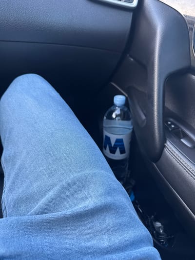 The image shows a close-up view of a person's legs wearing blue jeans, sitting in the passenger seat of a vehicle. A partially visible bottle of water with a blue "M" logo is placed in the door pocket. The interior of the car, including the dashboard and door panel, is visible. The scene is likely set in a residential area in Cojușna, Moldova, given the context of the photo.