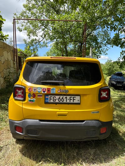 A yellow Jeep Renegade is parked on a grassy area, with trees and a stone wall in the background. The Jeep has several stickers on its rear windshield, including flags of various European countries. The license plate reads "FG-661-FF". A black SUV with a roof rack is visible in the background. The scene appears to be a rural area in the city of Navdaraantkari, Georgia.