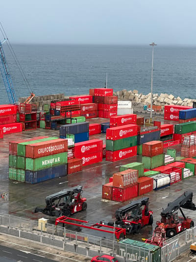 The image depicts a shipping container port in Puerto del Rosario, Fuerteventura, Spain, viewed from an elevated perspective. The scene is dominated by numerous multicolored shipping containers, predominantly red, with many bearing "BOLUDA LINES" branding. Other containers are green, blue, brown, and white, with some green containers displaying "GRUPO BOLUDA" and "NENUFAR." These containers are stacked multiple units high across the wet, dark grey paved ground of the port, suggesting recent rain.

In the foreground and midground, three large red container handling vehicles (reach stackers) are visible. Two face left, and one faces right, with one handler featuring an extended red frame attachment. A red passenger car is partially visible in the bottom foreground, parked near a metal barrier.

Towards the left, a blue gantry crane arm extends upwards. Along the ocean's edge, large grey concrete blocks, possibly tetrapods, form a protective barrier. The ocean itself is a dark, choppy grey under an overcast sky, meeting the land behind the container yard. A tall, multi-lamp light pole stands prominently on the right side of the image. In the distance near the concrete blocks, a white car and an orange utility vehicle are visible. Two individuals wearing high-visibility vests are present on the far left side, near a container stack. Small figures are also discernible on the platform of the tall light pole.