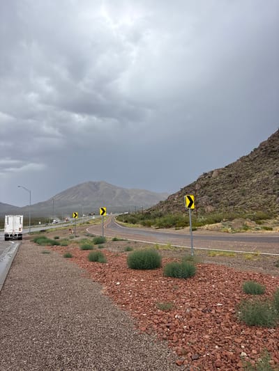 An overcast day captures a highway curving through a mountainous, arid landscape. In the foreground, a gravel shoulder transitions from dark gray to reddish-brown. A white semi-trailer truck is parked on the left shoulder. Yellow chevron signs indicating a curve are visible along the highway. Further in the distance, mountains with sparse vegetation rise against a cloudy sky. The road appears wet, suggesting recent rain. The location is identified as Etholen, United States.