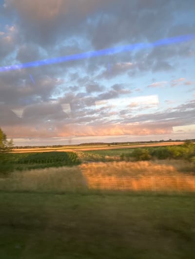 The image depicts a rural landscape at sunset, as viewed from a moving vehicle. The foreground is a blur of green grass. Behind this, a field of golden-brown crops, likely wheat or barley, is illuminated by the setting sun. A strip of darker green vegetation separates this field from a distant line of trees and more fields. The sky is filled with dramatic clouds, tinged with pink and orange from the sunset, and a bright blue streak, possibly a reflection or lens flare, cuts across the upper portion of the image. The environment suggests an agricultural area in the countryside. The location is specified as Mozów, Poland, though this cannot be visually confirmed from the image alone. There are no people or specific actions visible.