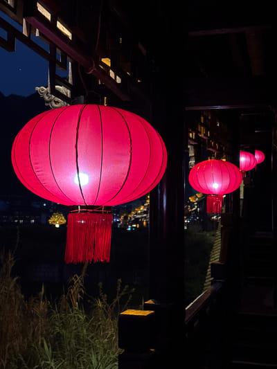 The image depicts a series of red lanterns hanging in Zhuoshui, China. The lanterns are spherical with horizontal stripes and tassels. The lanterns are illuminated from within and the background is dark, with the lanterns closer to the camera appearing larger and brighter. The lanterns are hanging from a dark wooden structure and are reflected in the water below. Other architectural details and vegetation are visible in the background.