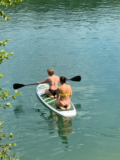 In Plintovka, Russia, two individuals are paddleboarding on a tranquil body of water under a bright sky. A woman with blonde hair, wearing a black and white patterned bikini top and black bottoms, is seated on the paddleboard, holding a black paddle. Beside her, a woman with dark hair pulled back in a bun, wearing a yellow bikini top and multi-colored striped bottoms, is kneeling on the paddleboard. The paddleboard is white with green and gray accents. The water is a clear, turquoise green, reflecting the sky and surrounding greenery. Foliage from trees drapes into the frame from the top left and lower left corners. The scene depicts a recreational activity on a calm lake or river.