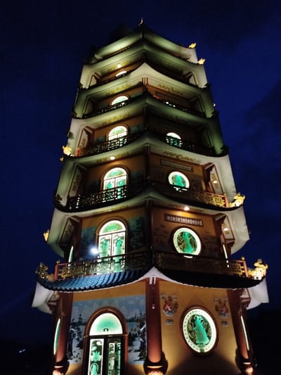The image displays a majestic, multi-tiered pagoda brightly illuminated at night, set against a dark, deep blue sky. Likely located in Phú An, Vietnam, the structure exhibits classic East Asian architectural elements with its upward-curving eaves, ornate balconies, and stacked rooflines.
The pagoda's facade is primarily a warm yellowish-orange, complemented by lighter cream or white structural details and a dark-tiled roof. Each level is adorned with intricate decorative elements, possibly depicting mythical creatures or traditional patterns, highlighted by warm artificial lighting. A striking feature is the array of windows—both arched and circular—which glow with a vibrant green light. Within these green-lit windows, silhouettes of what appear to be statues or figures, possibly Buddhist deities, are clearly visible, casting a serene and mystical aura. Dark, ornate railings line the balconies on each tier. The absence of daylight emphasizes the dramatic illumination of the architecture, creating a sense of grandeur and spiritual presence. No discernible text is visible in the image.