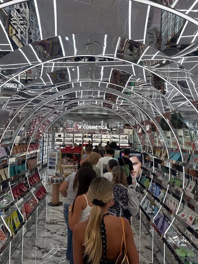 The image shows a queue of people inside a cosmetics store in Amadora, Portugal. The store has a futuristic design with a mirrored ceiling that reflects the brightly lit shelves of products. The shelves are stocked with various beauty products, including skincare, makeup, and fragrances. The floor is made of white marble with dark gray veins. The people in the queue are mostly women, and they are all facing towards the front of the store. The atmosphere appears to be busy and orderly.