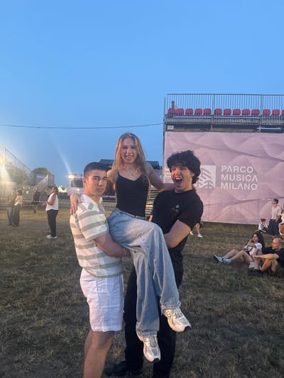 This media file is a photograph of three young people at an outdoor event in Segrate, Italy. The subjects are positioned in a playful stack, with one person at the bottom, another in the middle, and a third at the top. The person at the top is a man with dark, curly hair and a black t-shirt, who is leaning back and smiling broadly, looking at the camera. He is holding onto the person below him. The person in the middle is a woman with blonde hair, wearing a black top and jeans. She is smiling and looking at the camera, being supported by the person below. The person at the bottom is a man wearing a striped t-shirt and white shorts. He is holding up the other two people.
The scene appears to be a grassy field with a large banner in the background that reads "PARCO MUSICA MILANO." The banner is light purple with a wavy pattern. To the left of the banner, there are bleachers with red seats. The sky is clear and blue, suggesting it is daytime, likely late afternoon or early evening due to the lighting. Other people can be seen in the background, some sitting on the grass and others standing. The overall mood seems to be lighthearted and joyful, with the subjects engaging in a fun pose. The event appears to be a music festival or concert given the banner and the setting.