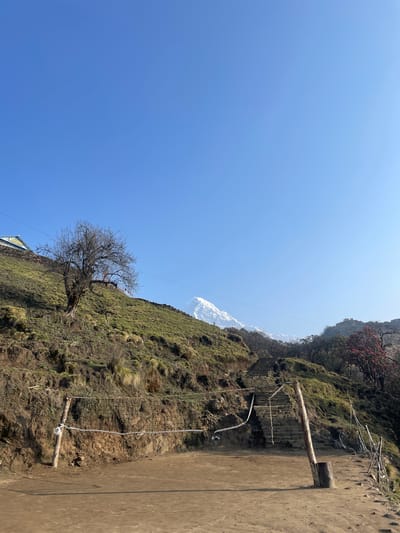 The image depicts a scene in Lwang Ghalel, Nepal. The landscape is characterized by a steep, grassy hillside with a bare tree in the foreground. The terrain slopes upward, revealing a distant snow-capped mountain peak against a clear blue sky. A rudimentary path runs along the edge of the hillside, supported by wooden posts and rope netting, acting as a safety barrier. The scene suggests a trekking or hiking route in a mountainous region.