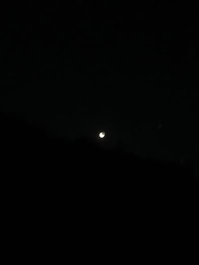 The image depicts a dark, night sky with a crescent moon visible in the upper central portion. The moon is a bright sliver against the blackness. There is a faint, dark horizontal line in the lower third of the image, possibly representing a distant landscape or horizon. No specific details about the city of Dehqonbozor, Uzbekistan, can be inferred from the image.