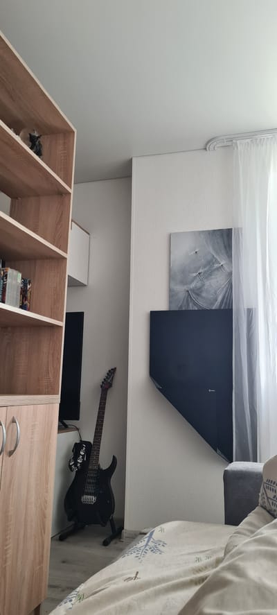The image depicts a room interior in Kohtla-Järve linn, Estonia. The room is furnished with light-colored wooden shelving units, some containing books and decorative items. Against the wall is a black electric guitar on a stand, a flat-screen television, and a white storage unit. Above the guitar, the wall is decorated with artwork; a dandelion print and a black abstract piece. The room has a low ceiling, and a curtain runs along the top of the wall. Part of a couch is visible in the upper right corner. No people are visible.