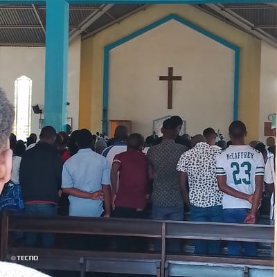 This image depicts the interior of a church in Machakos Central ward, Kenya, during a religious service or gathering. Approximately 20-30 individuals, predominantly adult males, are visible from behind, standing in pews and facing forward towards the altar area. The foreground features dark wooden pews. In the mid-ground, individuals are dressed in varied attire, including a white t-shirt with "McCAFFREY 23" printed on the back, a white shirt with black leopard print, and solid colored shirts (maroon, light blue, dark blue/black). Their hands are generally clasped behind their backs or at their sides.
The altar area features a large, dark wooden cross mounted centrally on a cream-colored wall, which is outlined in light blue with an inverted V-shape at the top. Below the cross is a grey rectangular panel. A person in green vestments is partially visible behind this panel. The church's interior architecture includes tall, light blue pillars, one prominently on the left. A tall, multi-paned window with an arched top is visible on the left wall, admitting natural light. The ceiling consists of exposed metal trusses and what appears to be a corrugated or paneled roof. The overall lighting is bright. A "© TECNO" watermark is visible in the bottom left corner of the image.