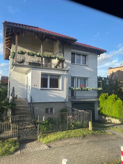 The image shows a two-story house with a red tile roof, located in Krosno Odrzańskie, Poland. The house is painted white and has a balcony on the upper floor with flower boxes filled with white flowers. There is a smaller balcony on the lower floor, also adorned with flower boxes. A set of concrete stairs leads to the entrance of the house, and a black metal fence encloses the property. A garage door is visible on the ground floor. The scene is set during the daytime, with a clear blue sky and sunlight casting shadows on the house and grounds. The surrounding area appears to be a residential neighborhood with some greenery and paved pathways.