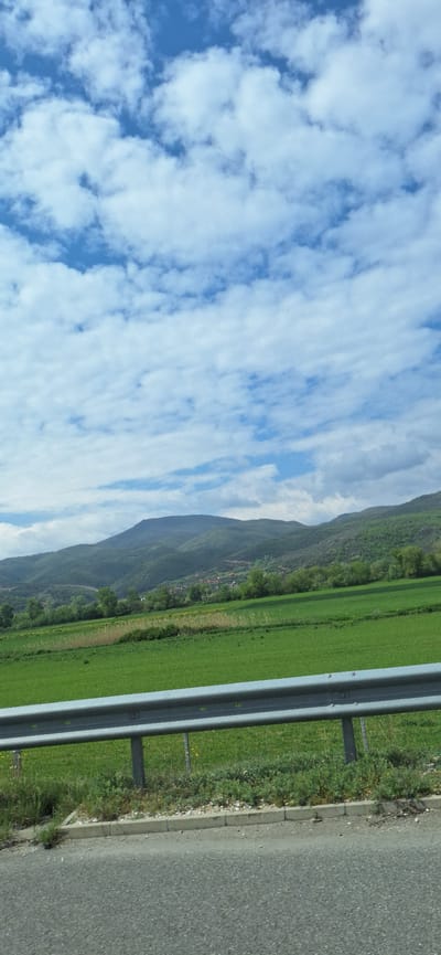 The image depicts a roadside scene, likely taken from a moving vehicle. On the left, a vibrant blue sky is filled with numerous white, puffy clouds. Adjacent to the sky is a mountain covered with trees and vegetation. A large green field, possibly farmland or a meadow, extends towards the right. A metal guardrail runs alongside the field, separating it from the road. A grey asphalt road occupies the rightmost section of the image, with a narrow strip of grass and gravel between the guardrail and the road. There are no people visible in the scene. The location could be Valkovo, Bulgaria, considering the landscape features and vegetation typically found in this region.
