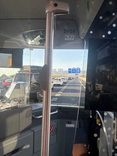 The image shows the interior of a bus looking forward through the windshield. The bus is in traffic on a highway, likely approaching or within Sant Boi de Llobregat, Spain, as indicated by road signs for Barcelona and other local areas. The scene is dominated by a line of cars and SUVs stuck in traffic. The bus has a metal pole running vertically in the center of the frame. On the dashboard of the bus, there is a ticket machine and a clear plastic barrier. Overhead, there are signs indicating "3804" and accessibility symbols (wheelchair, person). Reflections are visible on the windshield and interior surfaces. The overall impression is of a standstill on a sunny day, likely during commuting hours or a holiday period.