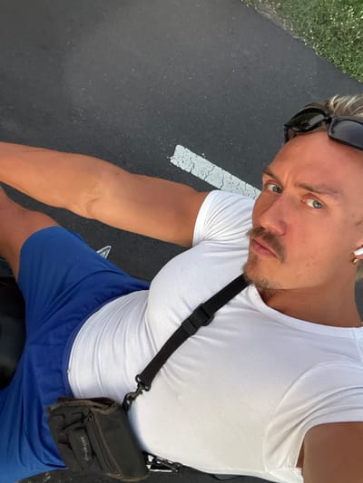 A man with blonde hair and blue eyes, wearing a white ribbed tank top and blue athletic shorts, poses outdoors on asphalt with a white stripe, seemingly in daylight. Sunglasses are perched on his head. He wears an earpiece in one ear and a black crossbody bag. The location is consistent with an outdoor road or path.