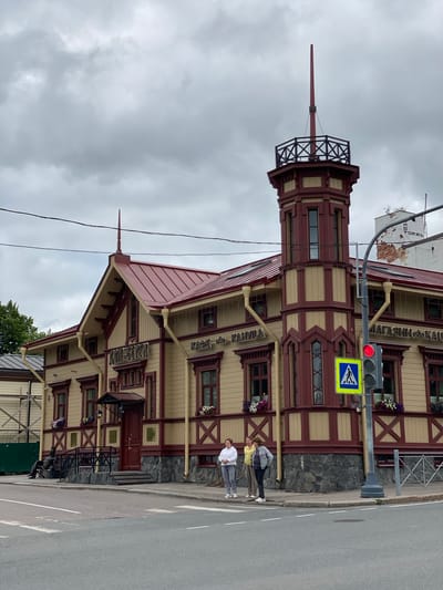 The image captures a street view of the historic "Dom Pervoy Svetopisi" (House of the First Painting) building in Sortavala, Russia. The building is distinguished by its red-and-yellow wooden architecture, featuring a prominent tower with a spire. The facade displays signage in Cyrillic, indicating "Dom Pervoy Svetopisi," "Cafe," and "Shop." Several people are visible: one person is seated on a bench near the entrance, and three individuals are standing on the sidewalk near a pedestrian crossing and traffic light. The sky is overcast, suggesting a cloudy day. The scene includes a traffic light, a pedestrian crossing sign, and a street intersection, indicating an urban setting.