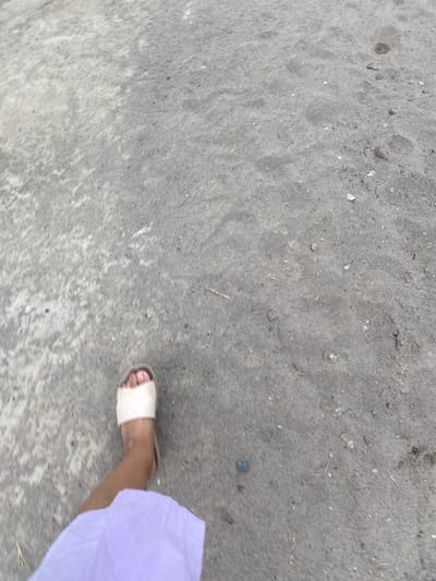 The image shows a person's right foot, wearing a cream-colored sandal, stepping on a rough, grey surface that appears to be a mixture of sand and gravel, possibly a dirt road or unpaved area. The person is wearing a light purple garment that extends from the knee down. The ground shows some small pebbles and debris. The context provided, indicating the location as the city of Kirov in Kyrgyzstan, suggests this is an outdoor urban or semi-urban environment. The image is taken from a high-angle perspective, looking down at the foot and the ground. There is motion blur, indicating the person is in motion, likely walking.