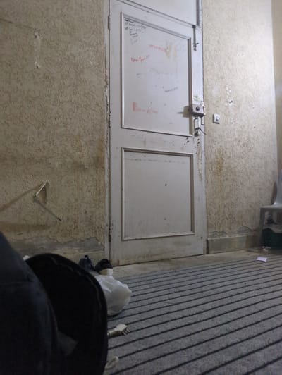 This image depicts a hallway or room with a textured, light-colored wall and a cream-colored double door. The door has a metal latch and what appears to be graffiti or writing in red ink on its surface. On the wall above the door, there's a light switch. To the right of the door, the floor is covered with a grey carpet featuring a striped pattern, and there are some objects on the floor including a white plastic bag, what looks like an electrical plug, and a black hooded garment in the foreground, suggesting a person is present but not fully visible. The overall scene appears to be indoors, possibly a residential or institutional setting, given the simplicity of the decor and the presence of functional elements like a light switch. The lighting suggests it could be daytime or artificial lighting is in use. There is no indication of weather conditions or specific time of day beyond the general interior setting. Visible text on the door includes names or messages written in red ink, but they are mostly illegible. The texture of the wall is rough and uneven, with some areas showing discoloration or dirt. The scene has a somewhat utilitarian and aged feel.