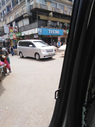 The image displays a street scene within Kikuyu Township ward, Kenya, captured from the interior of a vehicle looking forward and left. A silver Toyota Noah minivan, bearing license plate KCM 941C, is positioned on a paved asphalt road in the central foreground, angled slightly to the right. In the background, multi-story commercial buildings line the street. A prominent blue sign for "TECNO" is affixed to the first floor of a building, beneath signs for "TAI'S Bale Saloon." Other visible business signage includes "POSH SALON" and "CHEMIST," alongside a red "No Entry" traffic sign. On the left side of the frame, a person wearing a striped shirt and a green apron stands near a market stall, handling items in a red bag. Several other pedestrians are present on the sidewalks and street throughout the background. The immediate right foreground is partially obscured by the dark interior trim of the viewing vehicle, revealing a door frame and a metal latch. The street surface exhibits minor scattered debris.