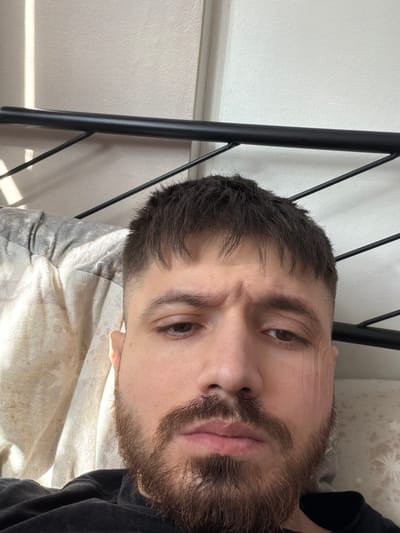 The image depicts a man with brown hair and a beard lying on a bed with a patterned sheet and pillow. He is looking directly at the camera with a neutral expression. A black metal bed frame is visible to the left of the frame. A light-colored wall is behind the bed, partially obscured by the bed frame. The location is not discernible from the provided image.