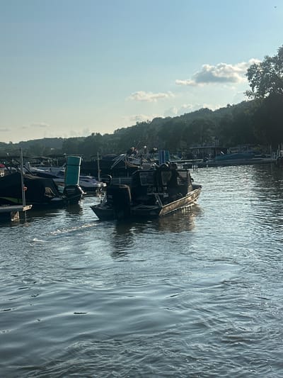 In Algonquin, United States of America, on a bright, clear day, a dark, flat-bottomed boat with multiple engines is moving across the water. The boat has a military or tactical appearance, with dark canvas covers and what appear to be military personnel on board. Several other boats are docked in the background, some covered with tarps. The water is rippled, with sunlight glinting off the surface. Trees and hills are visible in the distance, creating a scenic backdrop for the marina. The sky is a pale blue with a few scattered clouds.