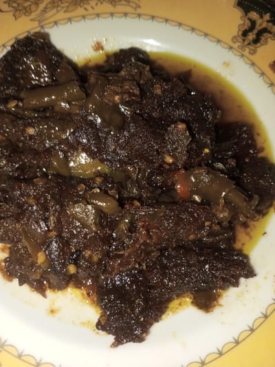 A dish containing dark brown, viscous food is presented in a white ceramic plate. The food consists of irregularly shaped, dark brown components mixed with fragmented green chili peppers and visible pale seeds, all suspended in a yellowish-brown oily sauce. The plate features a decorative rim with a yellow band adorned with white scalloped edges and dark brown ornamental patterns. This scene is located in Doramamidi, India.
