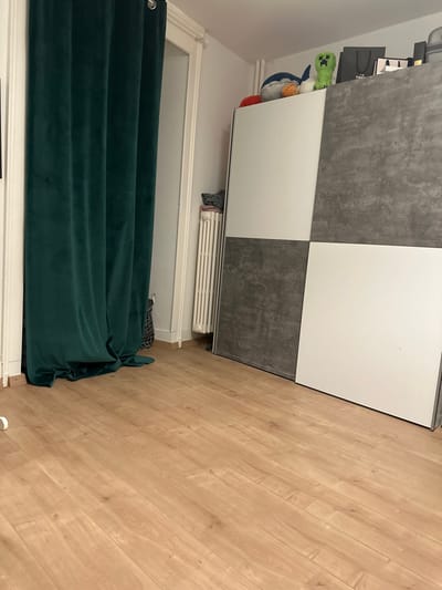 This interior photograph depicts a static scene within a room located in Montreux, Switzerland. The foreground is comprised of light wood laminate flooring. On the left, a dark emerald green velvet curtain with visible silver grommets hangs from a rod, obscuring a doorway or alcove, with its lower portion resting on the floor. A white doorframe is visible surrounding the curtain. To the right of the curtain, against a white wall, stands a white vertical panel radiator. A stack of folded gray fabric is positioned on top of the radiator. The right side of the frame is occupied by a large wardrobe or cabinet featuring four distinct panels: the top left and bottom right panels are white, while the top right and bottom left panels display a dark gray concrete-like texture. The top surface of this unit supports several objects, including a blue and white plush toy, an orange plush toy, a green pixelated plush toy, and multiple dark-colored paper shopping bags. No individuals are present within the frame, and no actions or interactions are occurring.