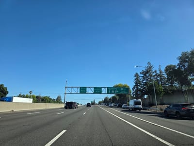 This media file captures a view from a vehicle traveling on a highway in Sacramento, United States of America. The scene is set under a clear, bright blue sky, indicating fair weather and likely daytime. The highway stretches ahead, with multiple lanes of asphalt marked by white lines. Several vehicles are visible on the road, including cars, vans, and trucks, suggesting moderate traffic.
Overhead, a large gantry displays green highway signs. The signs provide directional information, indicating exits and destinations. Text visible on the signs includes "80 EAST Reno," "EXIT 298 B Broadway 1/4 MILE," and "San Francisco WEST" along with "50 EAST South Lake Tahoe." These signs suggest the vehicle is on a route that connects to these major destinations.
On the right side of the highway, trees and a sound barrier line the roadside. On the left, a concrete barrier separates the lanes from what appears to be an embankment or industrial area, with a large white truck visible in the distance. The overall impression is a typical highway scene on a sunny day, with clear visibility and a sense of forward movement. The presence of the highway signs and the general landscape suggest an urban or suburban setting within the United States.