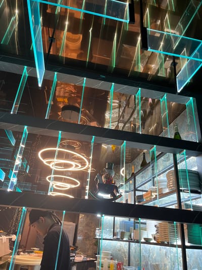 This image presents a modern, sophisticated interior, likely a bar, restaurant, or high-end retail space, characterized by prominent, illuminated shelving. The main feature consists of multiple vertical shelving units constructed with dark, possibly marble-clad or marbled-effect pillars supporting clear or frosted glass shelves. Each glass shelf is dramatically lit from beneath or within by vibrant blue-green LED lights, casting a cool, futuristic glow.

Various items are displayed on these shelves. Several wine or champagne bottles are visible, lying horizontally on different levels. Other shelves hold stacks of plates, some white and some green, along with other tableware. Further down, a teapot or kettle and what appears to be a small toy rocket ship can be seen.

The background is highly reflective, creating distorted reflections of the shelving, lights, and other elements of the room. A prominent feature in the mid-ground is a bright, warm-toned neon sign that clearly spells out "Coca".

On the far right side of the image, a person is partially visible, seen from the chest up. This individual, a male wearing a dark cap and a dark t-shirt, is looking towards the left, appearing to observe the shelving or the wider environment. His expression is neutral.

The strong artificial lighting, with the contrasting cool blue-green shelf lights and the warm "Coca" neon sign, suggests an evening or nighttime setting, or an interior designed for specific ambiance. The overall aesthetic is sleek and contemporary, fitting for a modern establishment in Basiglio, Italy. There is no specific activity taking place, beyond the presence of the individual in this stylish setting.