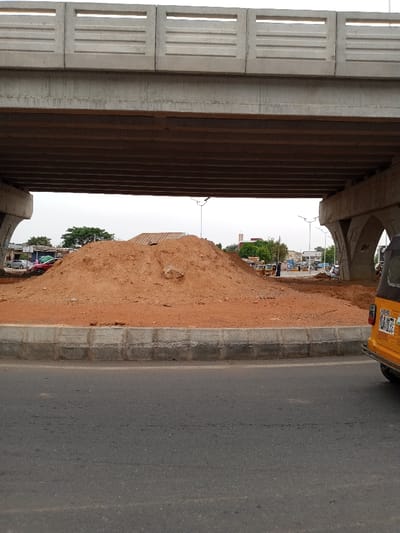 The image shows a large pile of dirt in the middle of a roundabout. The dirt pile is in front of a concrete bridge, which is located at the intersection of major roads. The roundabout is being constructed and there is a yellow three-wheeled vehicle parked on the right-hand side. There are several street lights and trees surrounding the roundabout.  The location is 9.279° N, 12.451° E, which is in Yola, Nigeria.