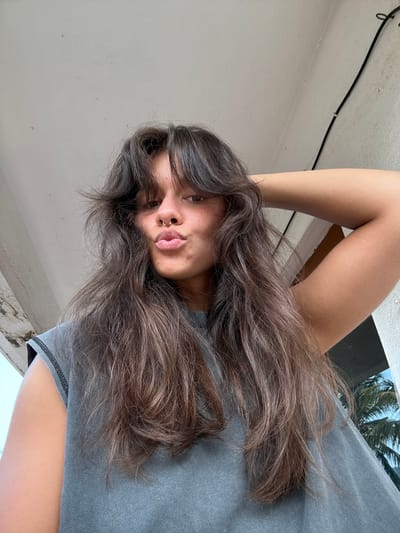 A woman with long, wavy brown hair and a septum piercing is facing the camera. She is wearing a grey shirt and has her arm raised behind her head. The background consists of a white wall, a black cable running along the wall, and a window showing a palm tree. The location cannot be determined as Marra, India, with the given information.