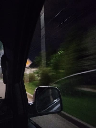 The image depicts a view from inside a vehicle at night, looking out the side window. The primary elements are the dark interior of the car, a side mirror reflecting the scene, and the blurred exterior passing by. The exterior shows a road with a grassy verge, trees and bushes on the right, and a building with a yellow sign in the distance on the left. A utility pole with wires is also visible. The motion blur indicates the vehicle is in transit. There is no indication of the city of Turkistan, Kazakhstan in the image.