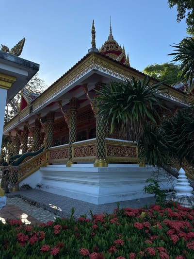 In Baan Tha Po, Thailand, a Buddhist temple's ornate facade is captured under a clear blue sky. The temple features a multi-tiered roof adorned with gold trim and decorative finials. Elaborate carvings and columns, painted in green and gold, support the structure. A wide, white staircase leads up to the temple entrance, bordered by sculpted Naga serpents. Lush green foliage and a bed of vibrant red flowers are situated in the foreground, adding a splash of color to the scene. The architectural style suggests a traditional Southeast Asian Buddhist temple.