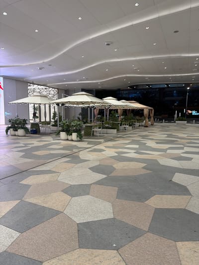 The image depicts an outdoor dining area in the Jianye District of China. The foreground features a geometric patterned tile floor in shades of gray, beige, and white. To the left, there are several tiered outdoor dining structures with white canopies, adorned with string lights. Potted plants in white containers are interspersed along the edges of the dining area, separating it from the pedestrian walkway. Tables and chairs are visible under the canopies, implying a restaurant or café setting.

The background shows a modern building with a curved, white ceiling featuring recessed lighting. A dark reflection in the glass walls hints at nighttime. A sign with an orange and white logo is visible on the building. The overall environment suggests a well-maintained, upscale outdoor dining space.