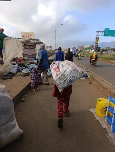 A child carries a large bag on their back along a roadside path. Several adults are nearby, and a smaller child stands next to an older man. A roadside stall with fabric and other goods is visible in the background. The setting is urban, with a highway running alongside the path, indicated by a road sign. The sky is overcast with clouds. A billboard advertises "Tushiike County" in the distance. A yellow container and a blue plastic shelf are next to the child.