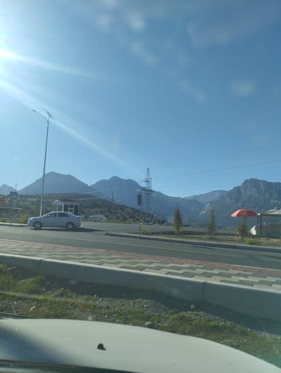 The image depicts a roadside view in Dardar, Uzbekistan, under a clear blue sky with prominent sun glare. In the foreground, the hood of a white vehicle is visible. The middle ground shows a paved road with a pedestrian walkway and a concrete curb. A silver sedan is parked on the side of the road, near a pedestrian crossing sign and a small white building. Beyond the road, a power pylon and several smaller trees are present. In the background, a range of imposing mountains forms the horizon, with some sparse vegetation on their lower slopes. A red umbrella is visible near a tent-like structure. The overall scene is one of a semi-urban or roadside environment with a backdrop of natural mountainous terrain.