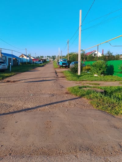 The image depicts a rural road in the city of Kranokholmskiy, Russia, on a clear day. The road is unpaved and has visible potholes and gravel. On the left side of the road, several houses with blue and red roofs are visible, along with parked cars. On the right side, a bright green fence lines the property, behind which lush green trees and vegetation can be seen. A tall utility pole with overhead wires runs along the right side of the road, as does a yellow gas pipe. Further down the road, a blue truck is parked, and another vehicle is partially visible. The sky is a clear, bright blue, indicating good weather.