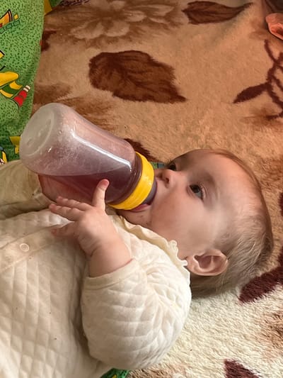 A baby is lying down on a surface of blankets and holding a baby bottle with a yellow nipple in their mouth. The bottle is filled with a purple liquid. The baby is wearing a cream-colored quilted shirt. The blanket beneath the baby is patterned with brown and cream colors. A second, green and multicolored blanket is visible in the lower left corner. The location in the city of Романята, Russia cannot be determined from the image.