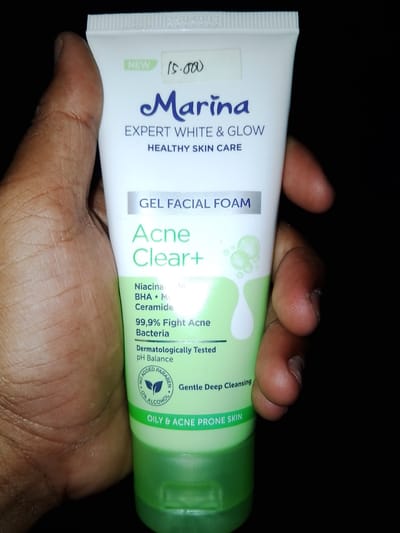 A white tube of Marina Expert White & Glow Gel Facial Foam for Acne Clear+ is held by a person's hand against a dark background. The tube has green accents and text indicating its purpose and ingredients. The image appears to be taken indoors. There is no discernible information in the image to suggest a location in Sendangrejo, Indonesia.