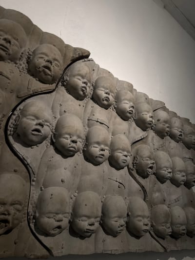 A close-up, slightly high-angle shot shows a gray, bas-relief wall sculpture comprised of multiple rows of rounded forms bearing stylized human faces. Each face is distinct, with open mouths and closed eyes, evoking a sense of repose or solemnity. The forms are arranged in an ascending, terraced pattern on a flat, rectangular surface. Small, raised spheres are scattered across the surface surrounding the faces. Thin, dark lines accentuate the borders of each row and the edges of the rectangular surface. The backdrop is a smooth, light gray wall. The sculpture is located in Epagny, Switzerland.