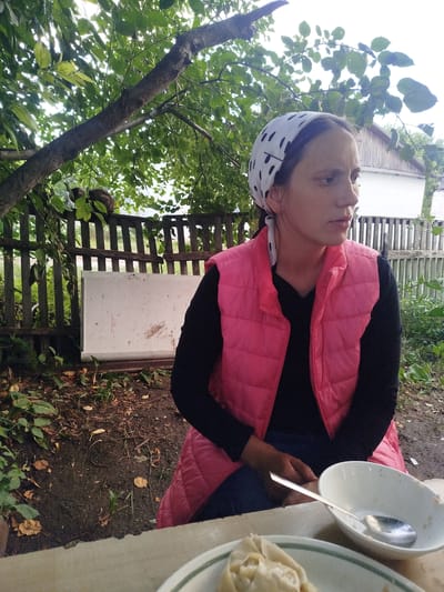 A young woman with a white bandana with black polka dots is sitting at a table outdoors. She is wearing a pink quilted vest over a black long-sleeved shirt and blue jeans. The woman is looking off to the right with a thoughtful expression. In the foreground, a white bowl with a silver spoon rests on the table, with some food visible in another bowl just below it.

The setting appears to be a rustic yard. A wooden fence runs across the background, with trees and foliage behind it. A white, rectangular object, possibly a cooler or a storage box, is leaning against the fence. A cat is perched on top of the fence, partially obscured by leaves. The ground is covered with dirt, fallen leaves, and some greenery. A house with a white exterior and a light-colored roof is visible in the distance. The lighting suggests it is daytime, possibly late afternoon, with a slightly overcast sky filtering through the trees. The overall mood is casual and peaceful, suggesting a moment of quiet contemplation during an outdoor meal. The location context indicates this scene is in Zarechnoye, Kazakhstan.