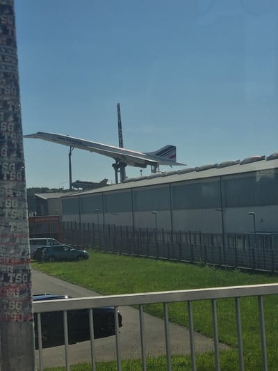 The image shows a Concorde supersonic passenger jet, specifically an Air France model, displayed at the Auto & Technik Museum Sinsheim in Sinsheim, Germany. The aircraft is positioned adjacent to a large, hangar-like building with several windows. The exterior also features the words "SB-WASCHEN" (self service carwash in German). A light blue car is parked near the carwash. A green lawn separates the museum building from a pedestrian walkway. Part of a fence with graffiti (with the letters "TSG") can be seen in the foreground, close to the viewer. The sky is clear.