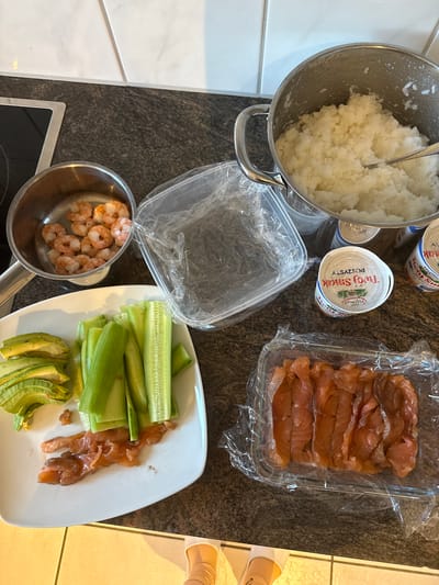 In Lufingen, Switzerland, this overhead shot captures a kitchen counter laden with ingredients for sushi. A pot of white rice sits to the right, with a spoon resting inside. To the left, a small metal pot contains cooked shrimp. A white plate holds sliced avocado, cucumber sticks, and pieces of smoked salmon. Another container, also with smoked salmon, is visible in the foreground. Several tubs of cream cheese are scattered around the scene, along with a clear plastic container and a roll of plastic wrap. The scene is set against a speckled countertop and a tiled backsplash.