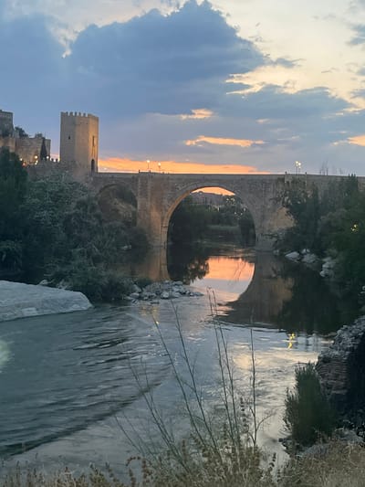 The image captures a serene sunset scene over the Tagus River in Toledo, Spain, with the historic Alcántara Bridge prominently featured. The sky is a canvas of soft blues and oranges, with scattered clouds reflecting the warm hues of the setting sun. The stone bridge, with its characteristic arches, spans the river, and its reflection is visible on the calm water. On the left bank, a fortified tower, part of the castle complex, stands sentinel against the twilight sky, its stonework illuminated by artificial lights. Lush greenery spills down the riverbanks, with trees and bushes adding texture to the landscape. The water itself is a dark, reflective surface, interrupted by the gentle flow of the river and the shapes of rocks and boulders. The overall atmosphere is one of peaceful antiquity, a timeless vista of a historic Spanish city.