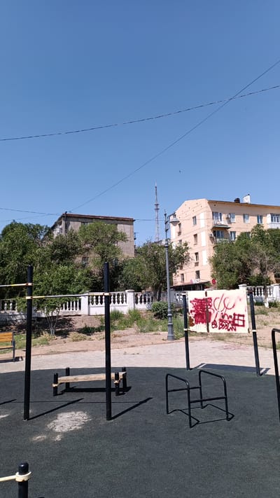 The image depicts an outdoor exercise area with various equipment, including parallel bars and a pull-up bar. The ground is covered with dark, textured rubber. In the background, there are trees, a white decorative fence, and buildings. One building is a light yellow, multi-story apartment building, while another is a darker, more industrial-looking structure. A radio tower is visible in the distance. A sign with red graffiti is also present. The sky is clear and blue. This scene is located in the city of Balqaş, Kazakhstan. There are no people or animals visible in the image.