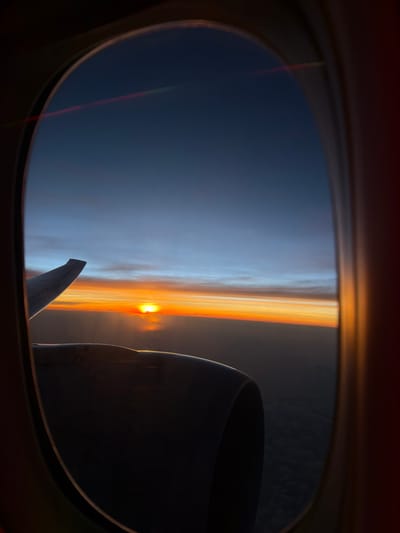 From an airplane window, a sunset horizon is seen, bisecting a blue sky above and a darker ground below. The wing and engine of the plane are partially visible. No people are seen. Location cannot be determined from this image.